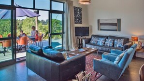 A modern living room with a sofa, armchairs, and a TV. Large windows offer a view of greenery outside. - The Paris Suite cottage - gorgeous views on 7 acres Mapua seaside village Nelson (Upper Moutere)