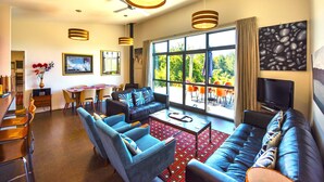 A modern living room with a sofa, chairs, a coffee table, and a TV. There is a dining area with a table and chairs, and a large window with a view of the outdoors. - The Paris Suite cottage - gorgeous views on 7 acres Mapua seaside village Nelson (Upper Moutere)