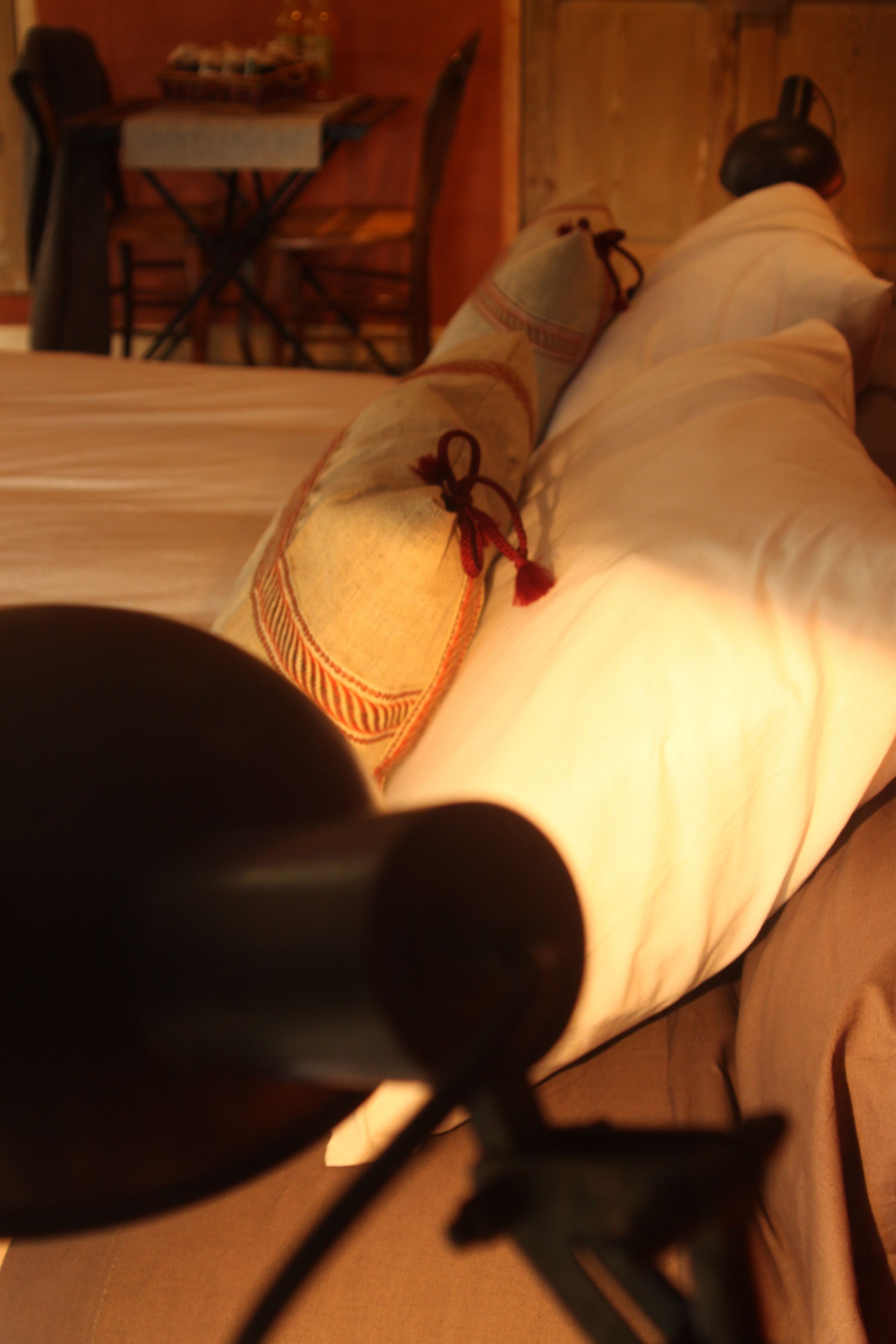 A bed with a pillow and a black object on it, possibly a lamp, in a room with a table and chairs in the background.