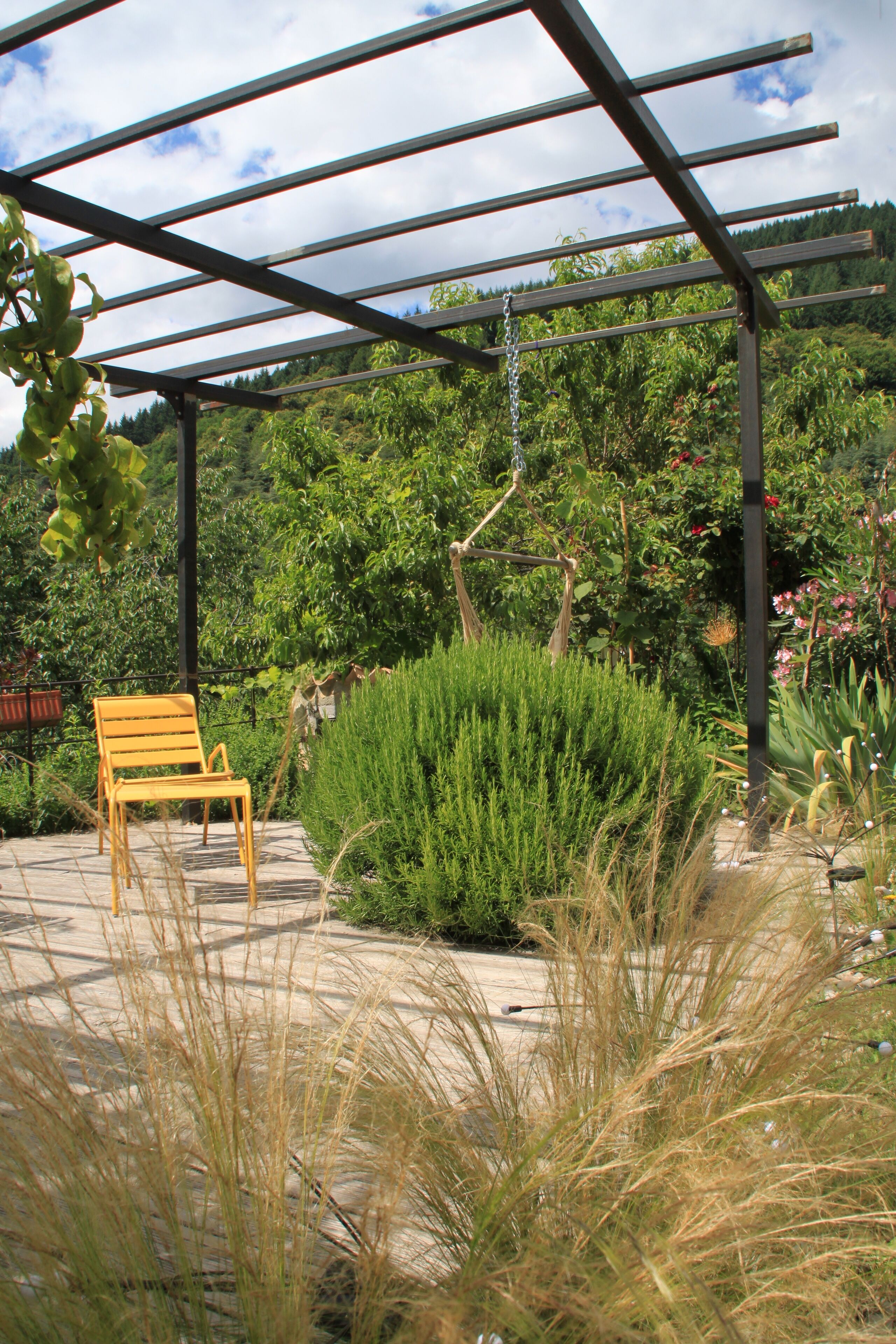 A covered outdoor seating area with a bench, surrounded by lush greenery and a pergola structure.