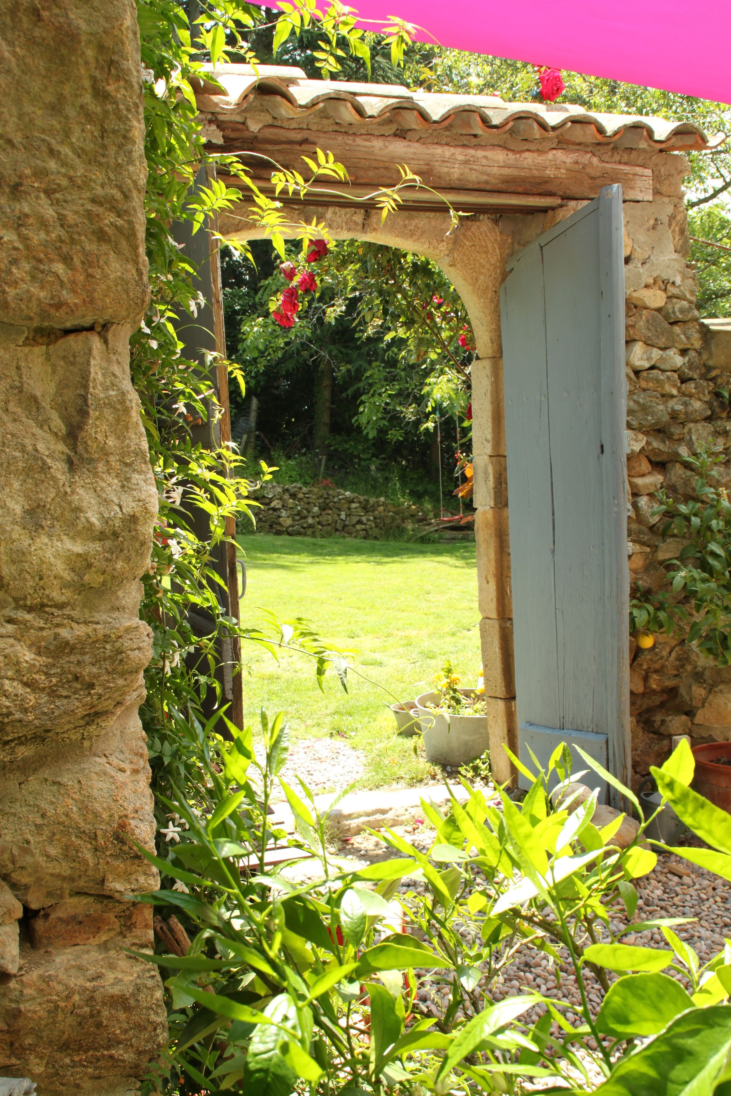 Arcada de pedra que dá acesso a um jardim com pérgola, plantas em vasos e um muro de pedra.