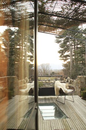 Una terraza de madera con piso de vidrio, rodeada de muros de piedra y macetas con plantas.