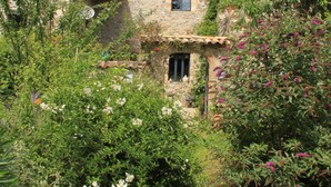 Una casa de piedra rústica con tejado de baldosas, rodeada de exuberante vegetación y plantas en flor.
