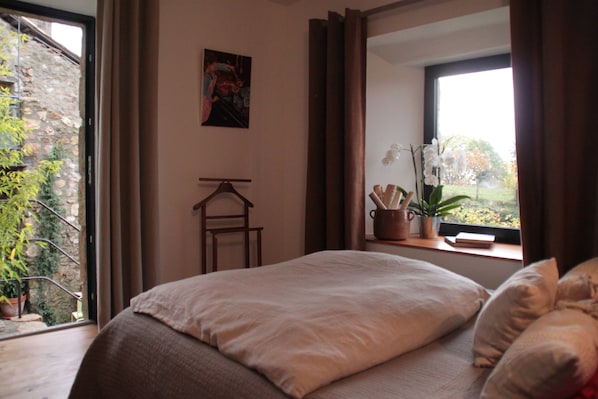 A bedroom with a bed, a window with curtains, a small table with a plant, and a wall-mounted picture. - La Demeure Des Maitres (Chirols)