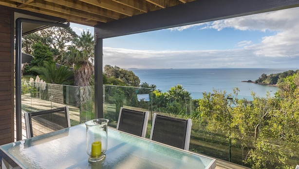 A glass-top table with chairs on a balcony overlooking a coastal view. - SPECTACULAR SEA VIEWS!! New Modern Apartment overlooking Bay.  (Oneroa)