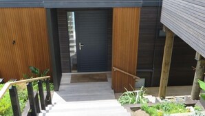 A modern house with a wooden exterior, a dark front door, and a staircase leading to a garden area with plants and a wooden deck. - SPECTACULAR SEA VIEWS!! New Modern Apartment overlooking Bay.  (Oneroa)