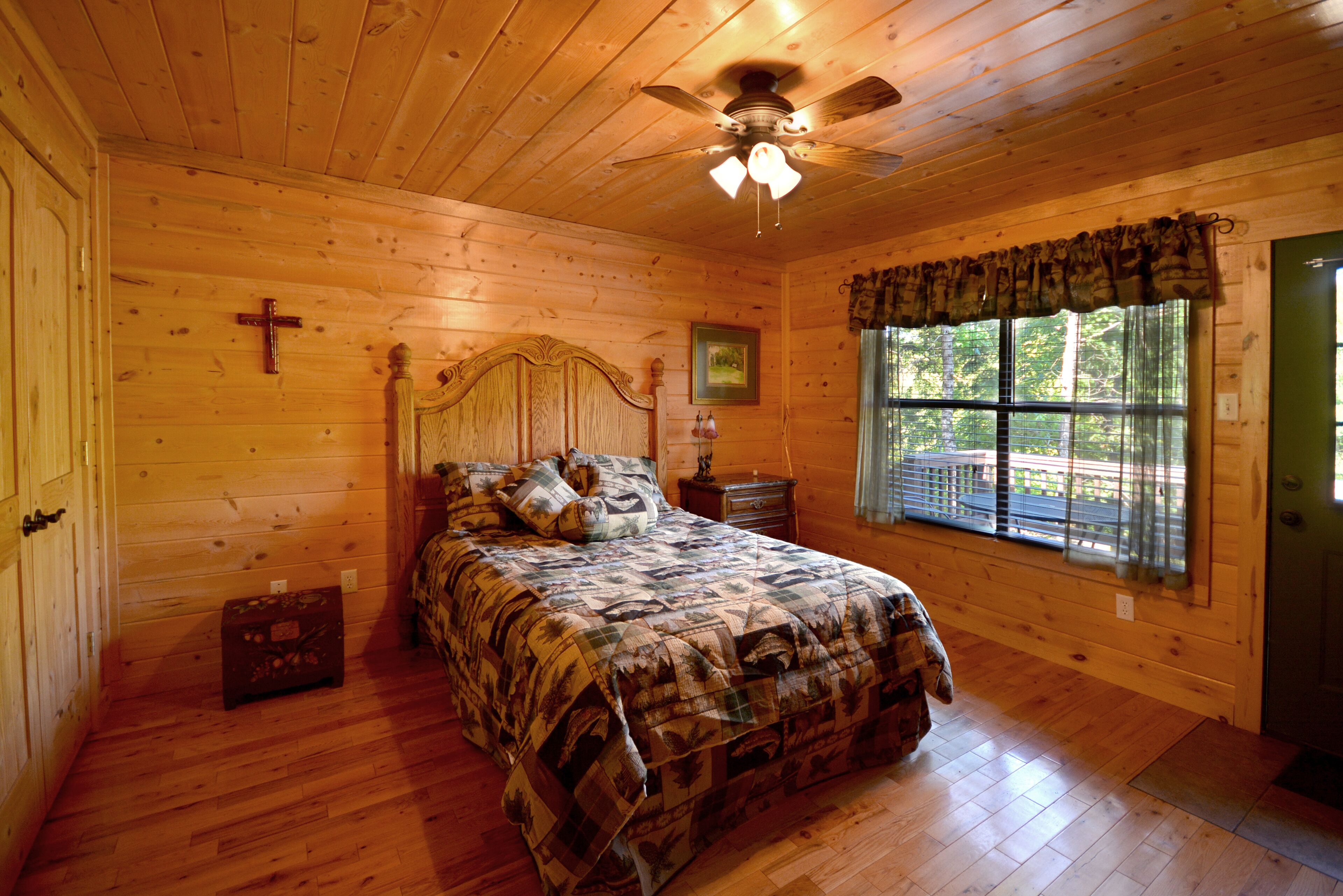 A wooden cabin bedroom with a bed, a cross on the wall, a ceiling fan, and a window with a view of the outdoors.