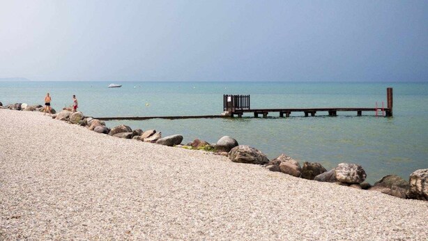 Ein Kiesstrand mit einer hölzernen Steg, der sich über ruhiges Wasser erstreckt, einem kleinen Holgebäude am Ende und einem wolkenlosen Himmel.
