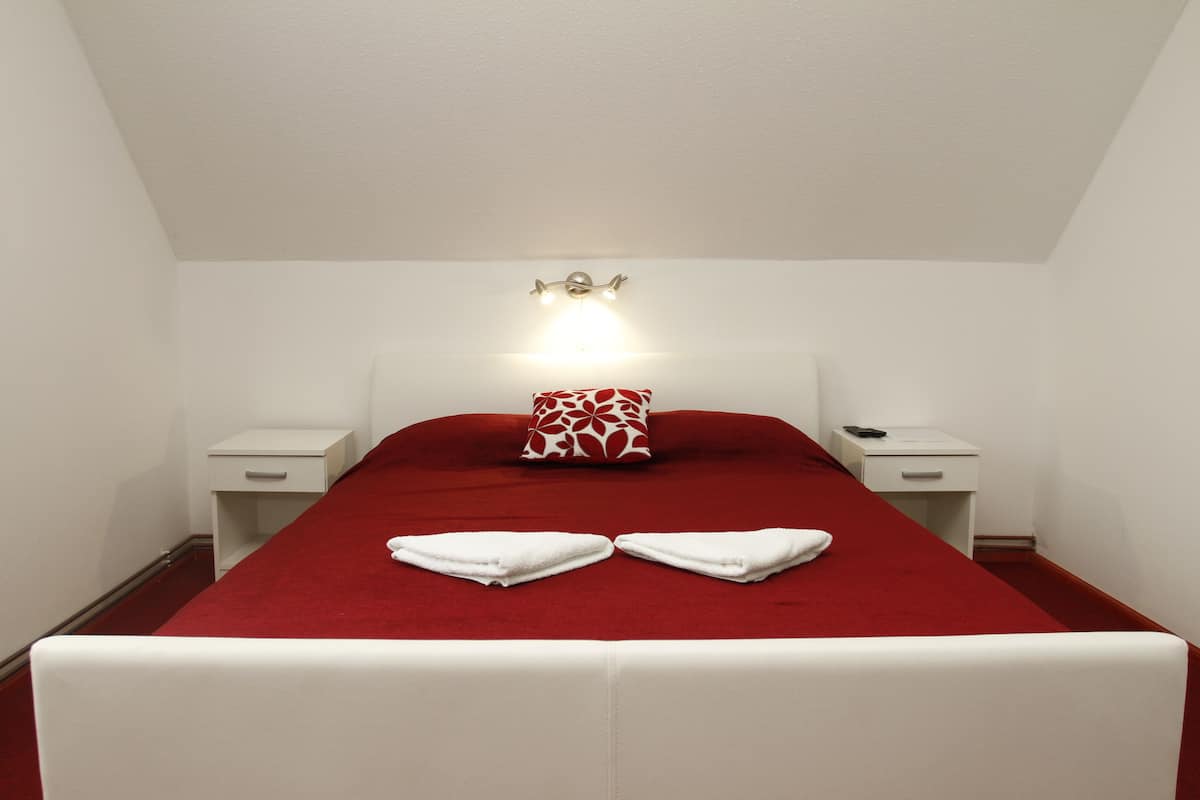 Ein Hotelzimmer mit rotem Bettbezug, weißem Kopfteil, zwei Nachttischen, einem roten und weißen Kissenmuster sowie einer wandmontierten Leuchte.