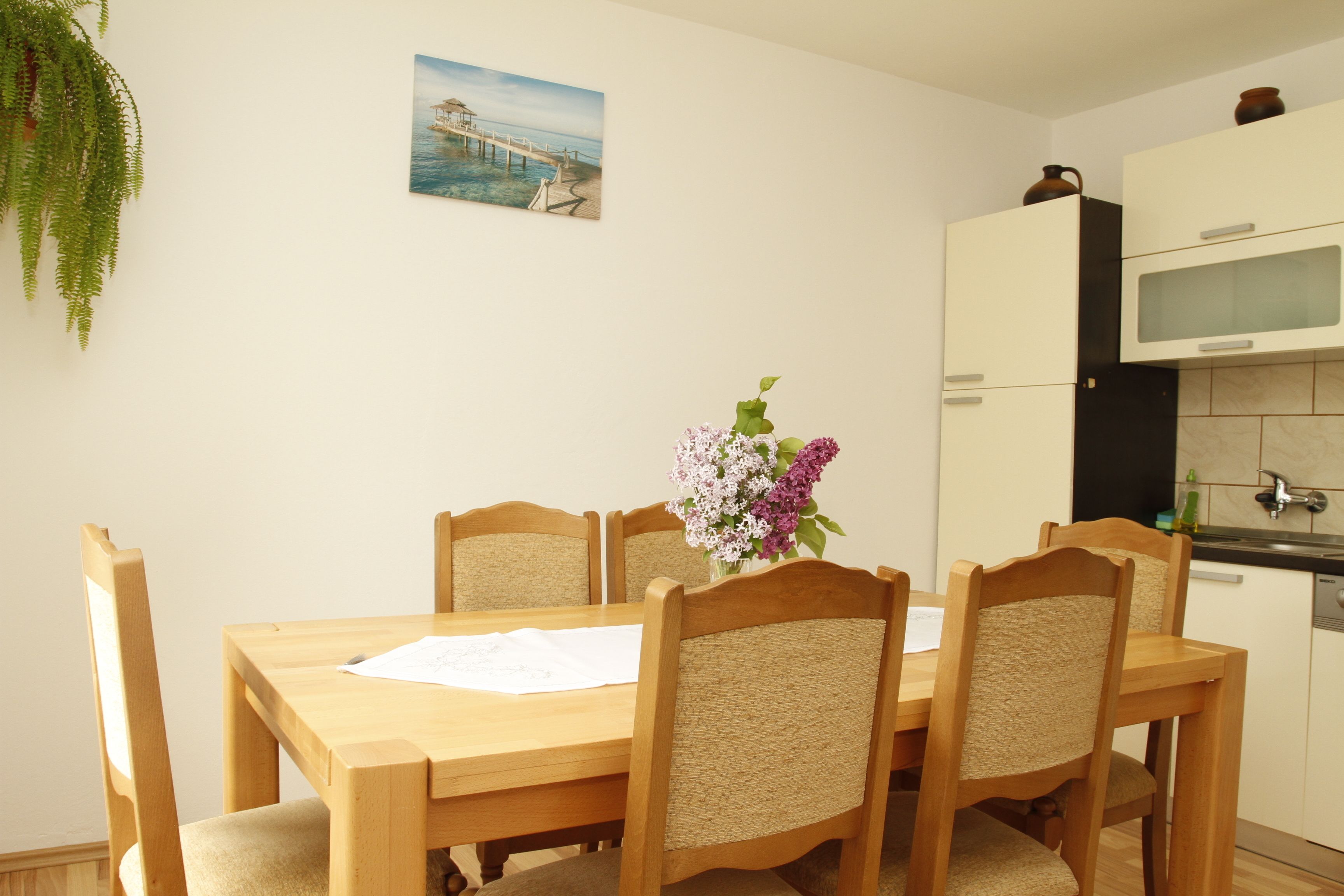 A dining area with a wooden table and chairs, a vase with flowers, and a kitchen in the background.