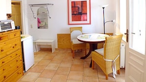 Une petite pièce bien éclairée, avec une table à manger, des chaises, un petit meuble blanc et un micro-ondes posé sur une commode.