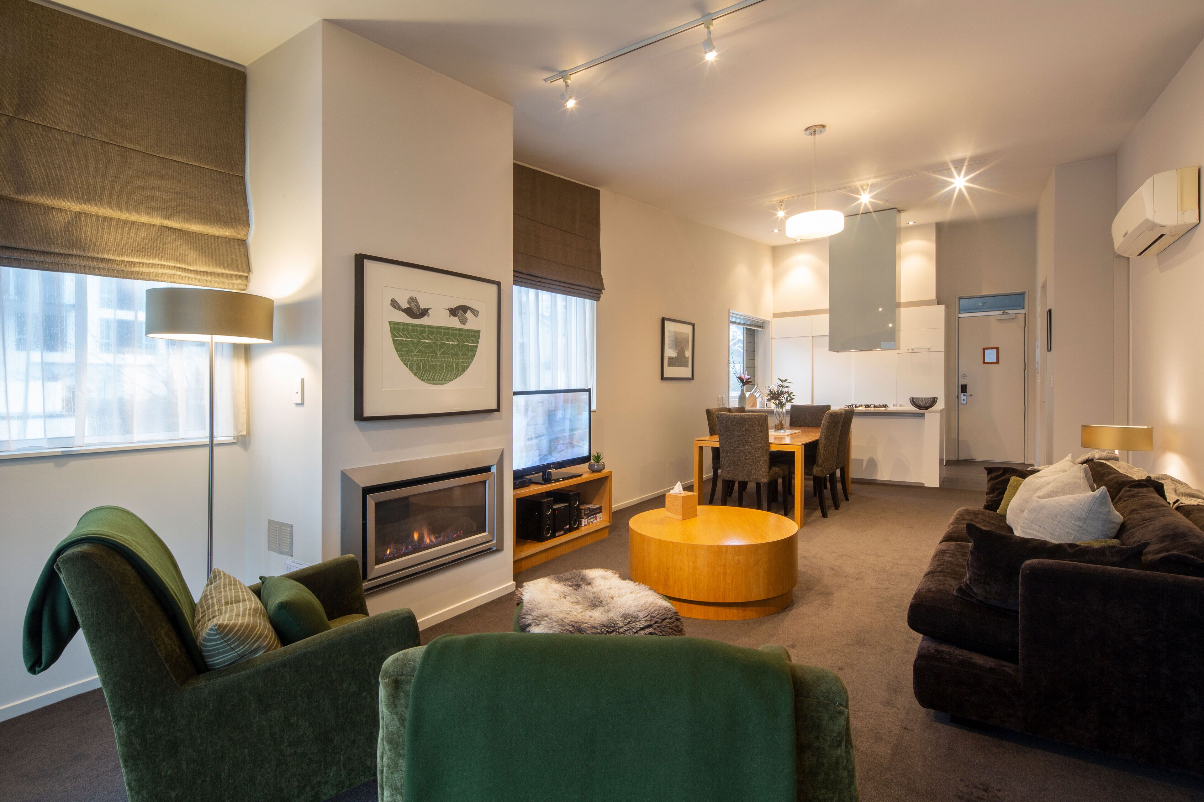 A modern living room with a fireplace, green armchairs, a dark sofa, and a dining area with a table and chairs.