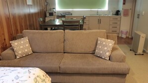 A cozy living room with a beige sofa, patterned cushions, a floral bedspread, and a dining area with a table and chairs.