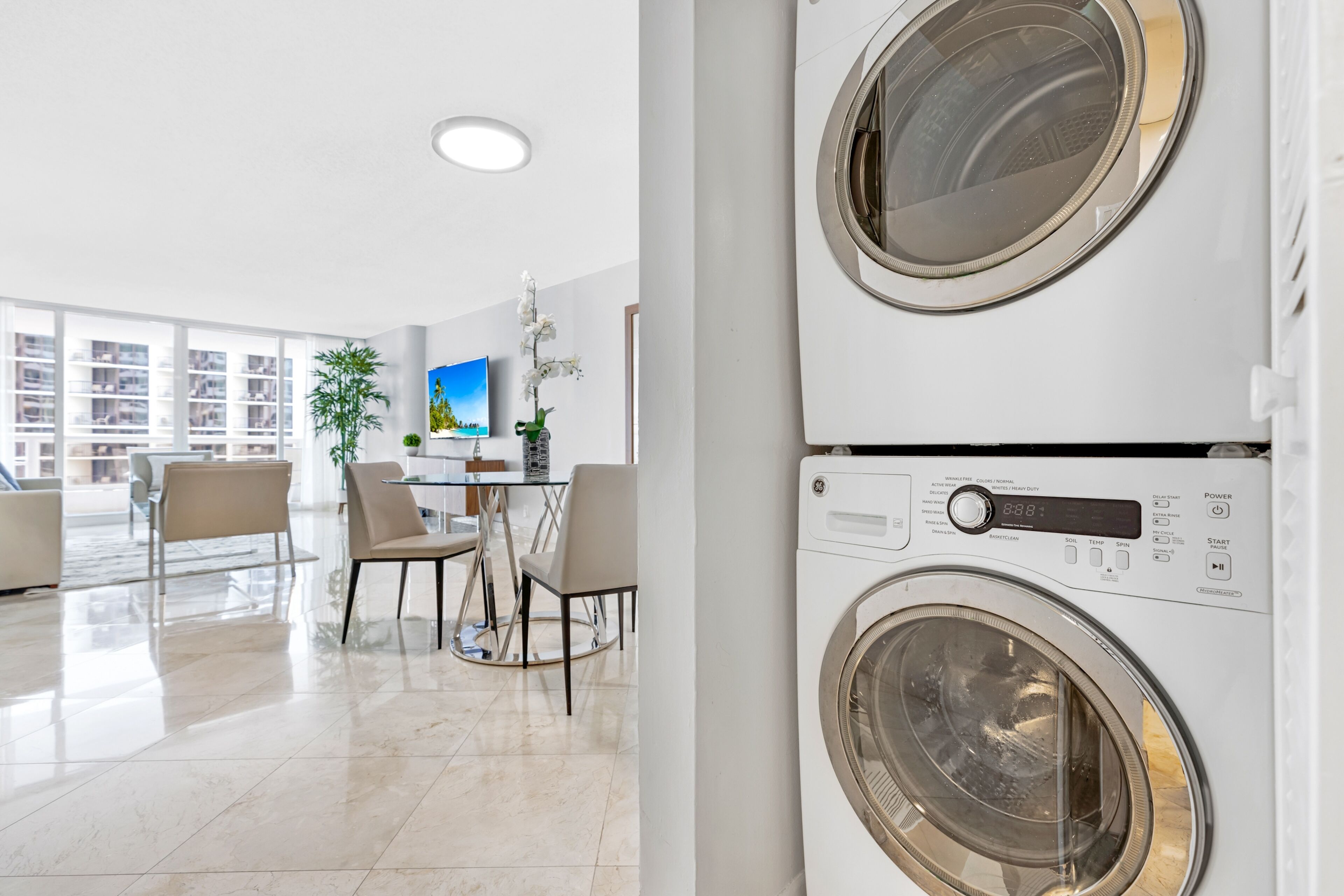 A modern apartment with a dining area, two stacked washing machines, and a large window offering a city view.