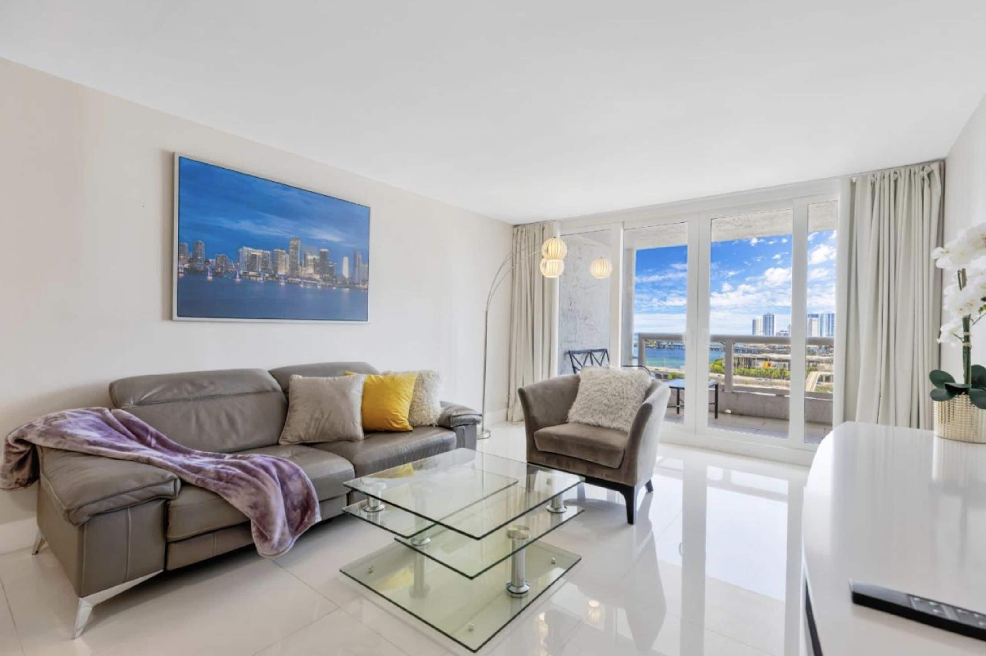 A modern living room with a glass coffee table, a sofa, and a chair. There is a large window with curtains, a painting of a cityscape, and a view of a bridge.