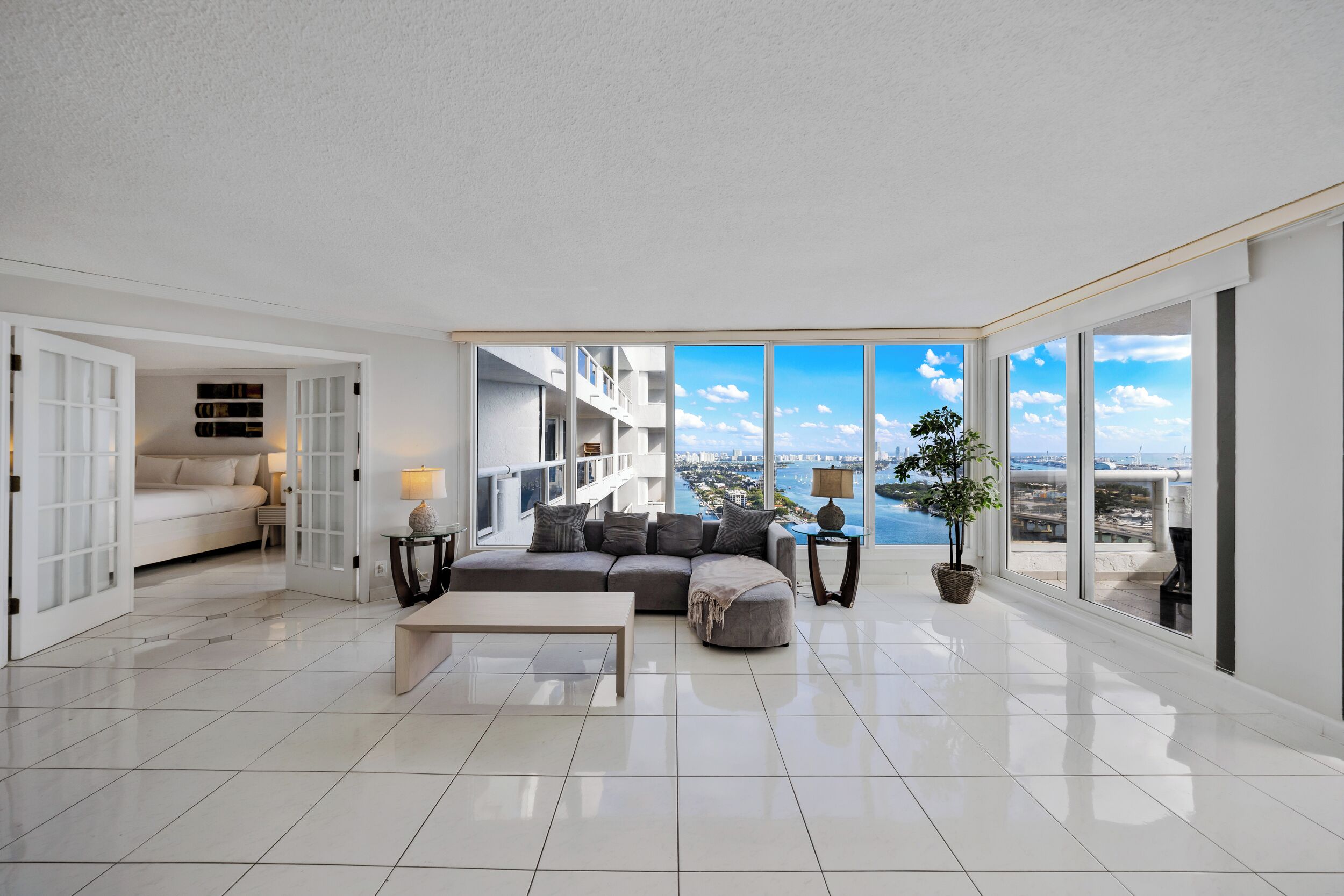 A spacious living room with a large window offering a city view, a grey sectional sofa, a coffee table, and a potted plant.
