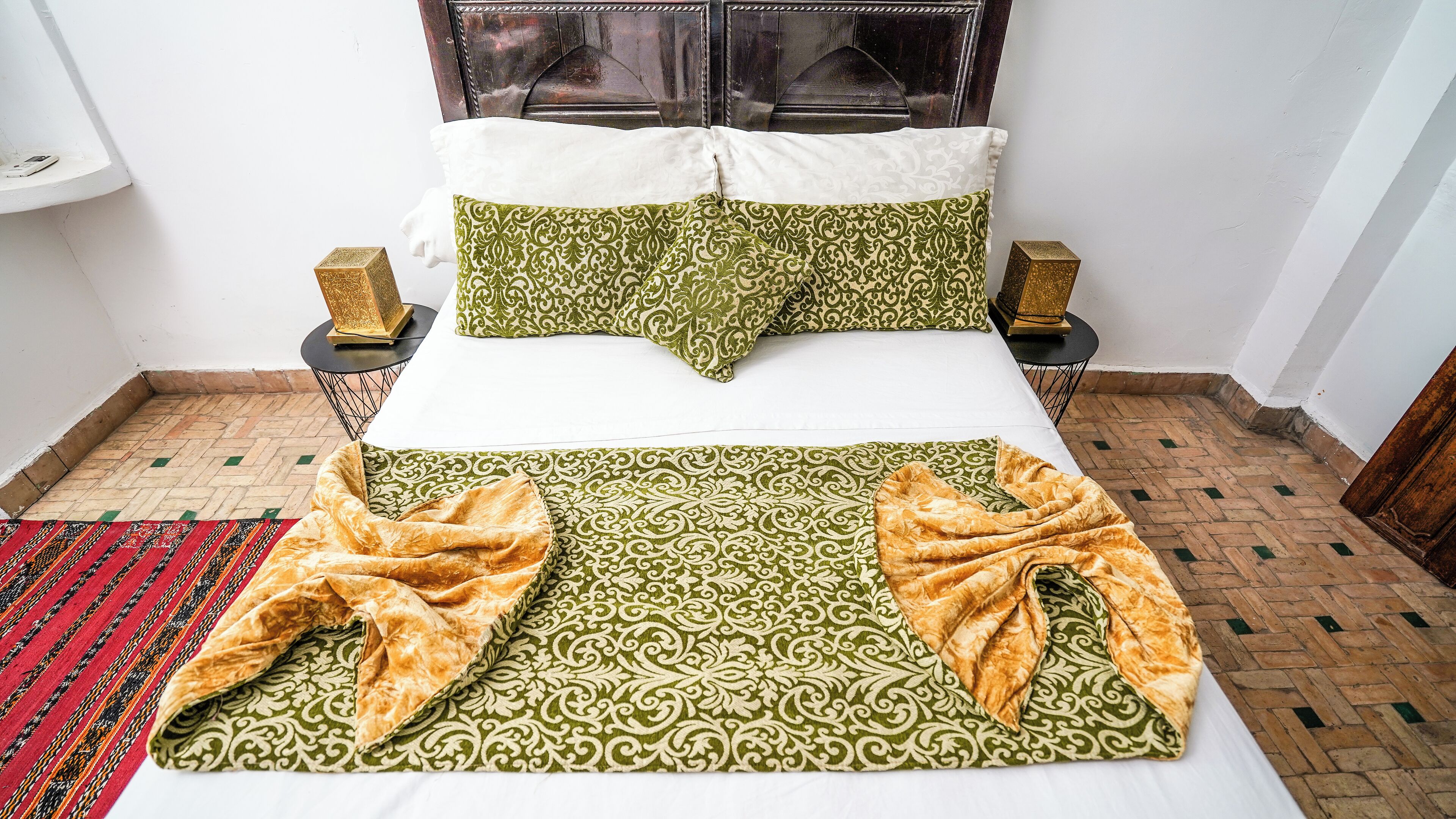 A neatly made bed with patterned pillows and a green and gold blanket, two bedside tables with decorative items, and a patterned rug on the floor.