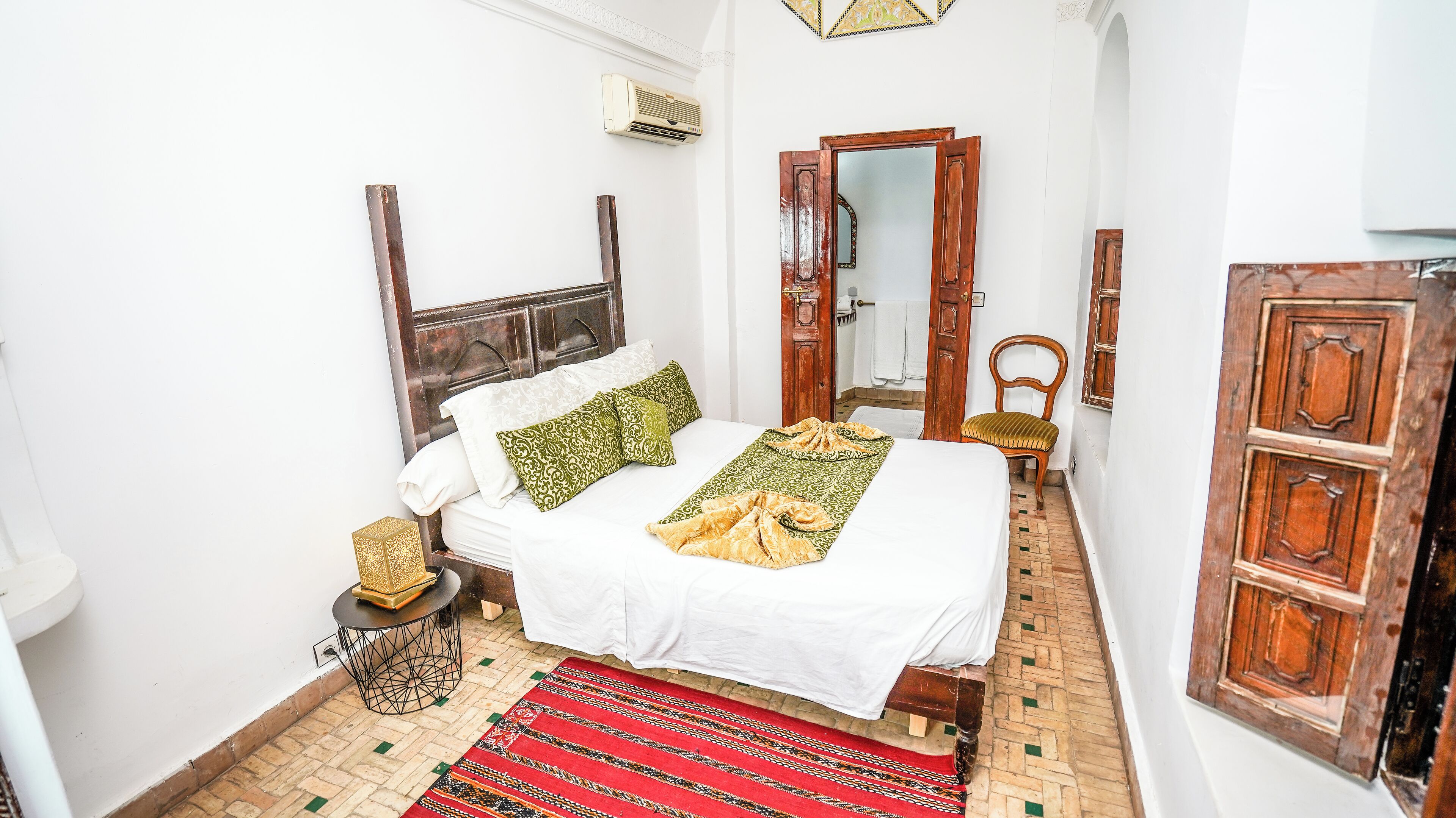 A narrow room with a bed, a chair, a small table, and a wooden door.