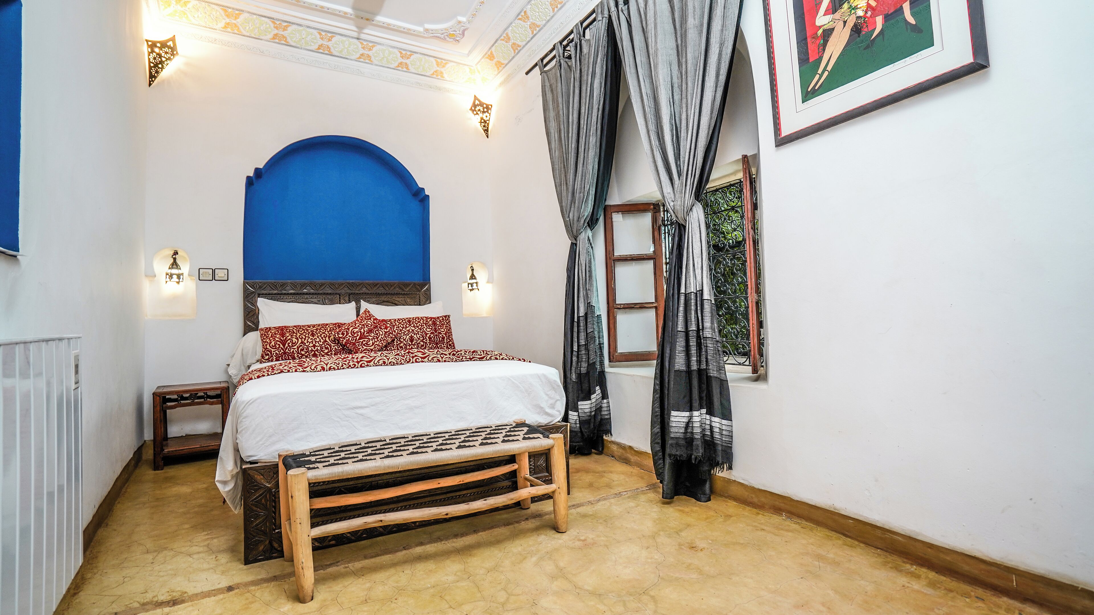 Un dormitorio con cama, un banco, una ventana con cortinas e iluminación en la pared.