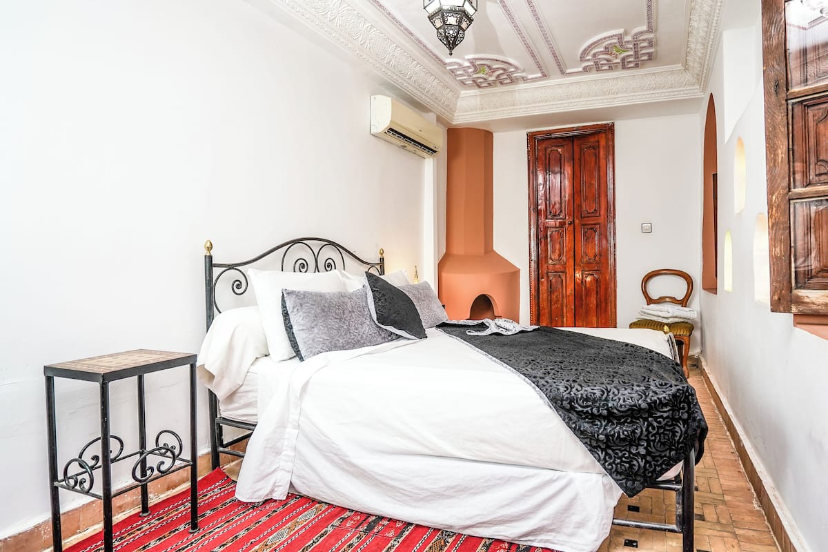 Un lit bien fait, avec une couverture et des oreillers à motifs, une petite table de chevet, un tapis rouge à motifs et une porte en bois, dans une pièce aux murs blancs et au plafond décoratif.