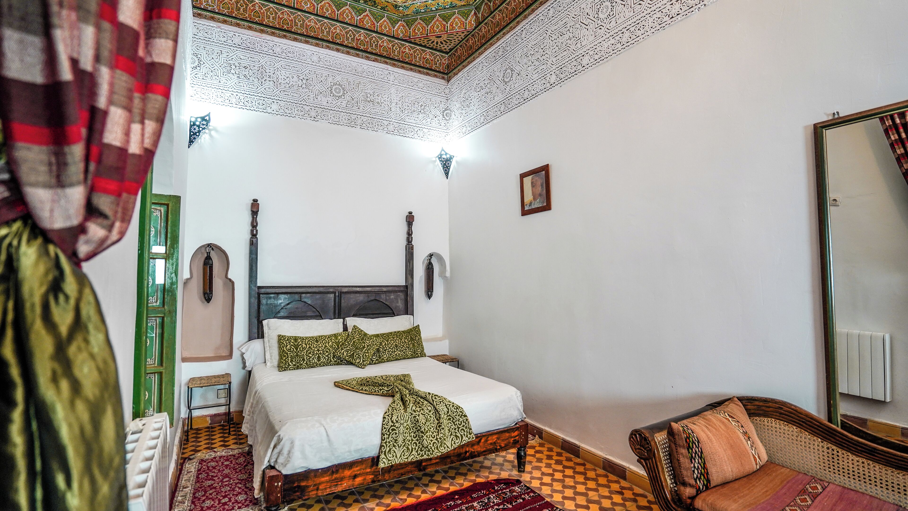 Una habitación tradicional con cama, cabecero de madera, alfombra estampada y espejo colgado en la pared.
