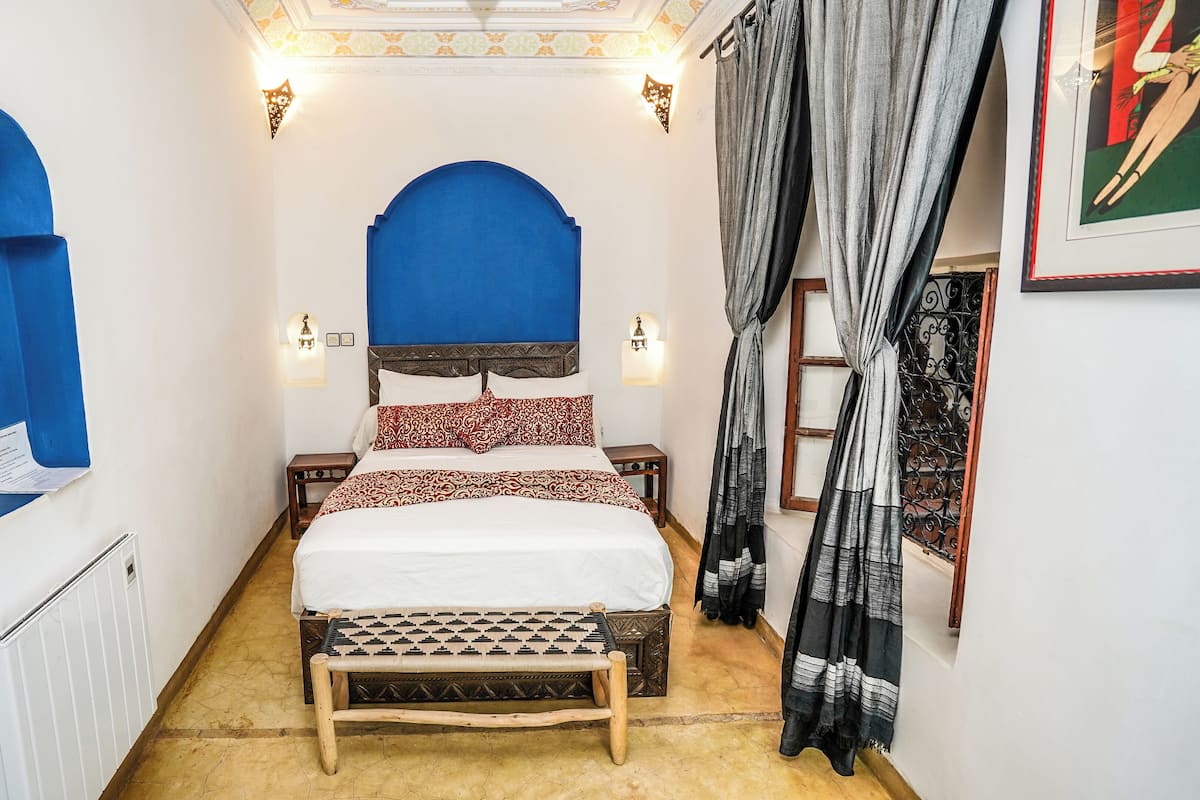 Une chambre d’hôtel étroite, avec un lit, une petite table et un banc.