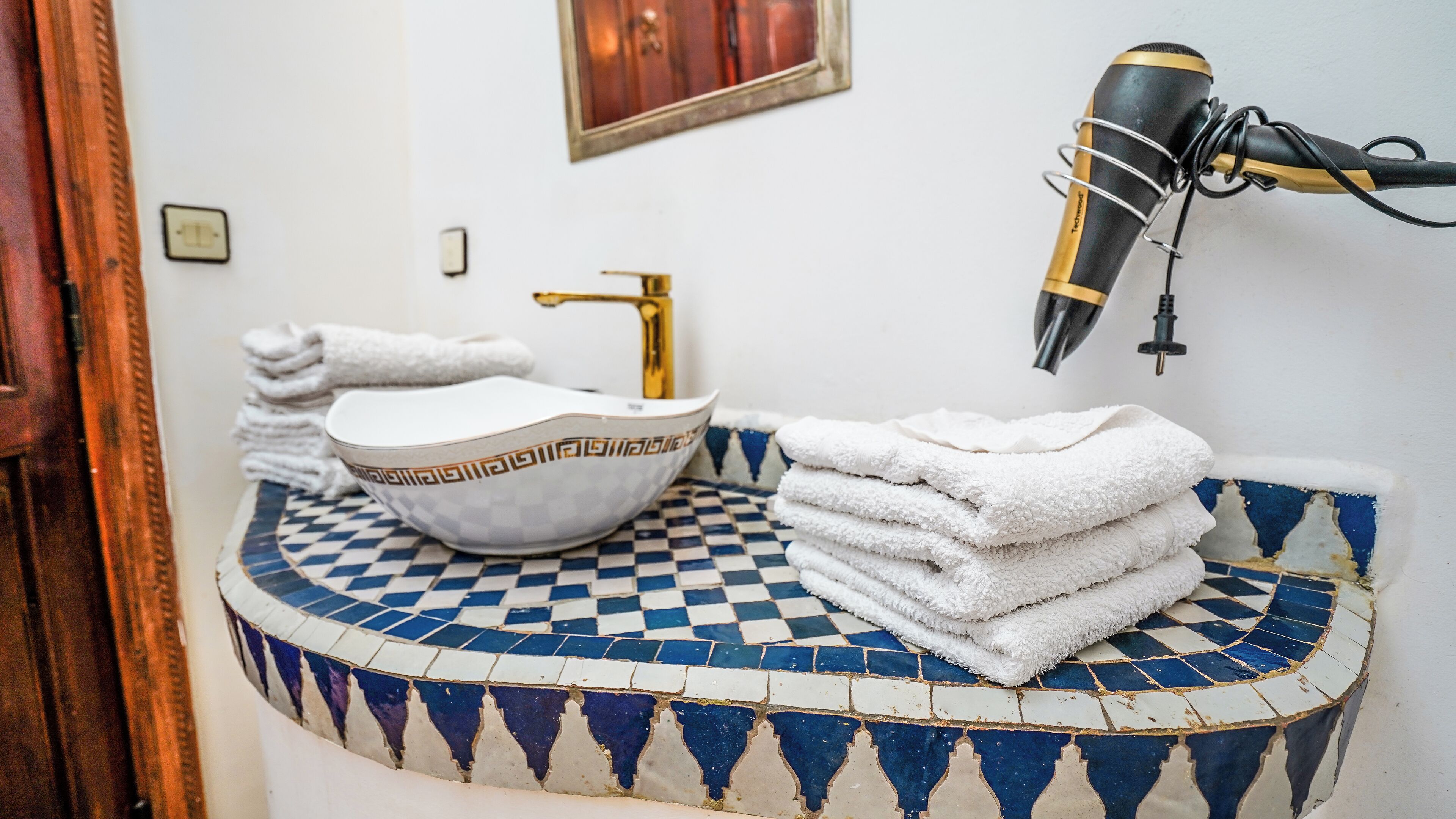 A bathroom with a checkered blue and white tiled floor, a white sink with a gold faucet, a white bowl, and a stack of white towels.