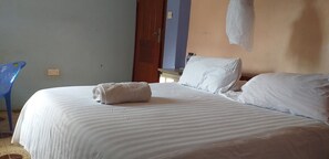 A single bed with a white striped bedspread and two pillows, a blue chair, and a wooden door in a room with light blue and beige walls. - Royal Pine Cottages (Ruiru)
