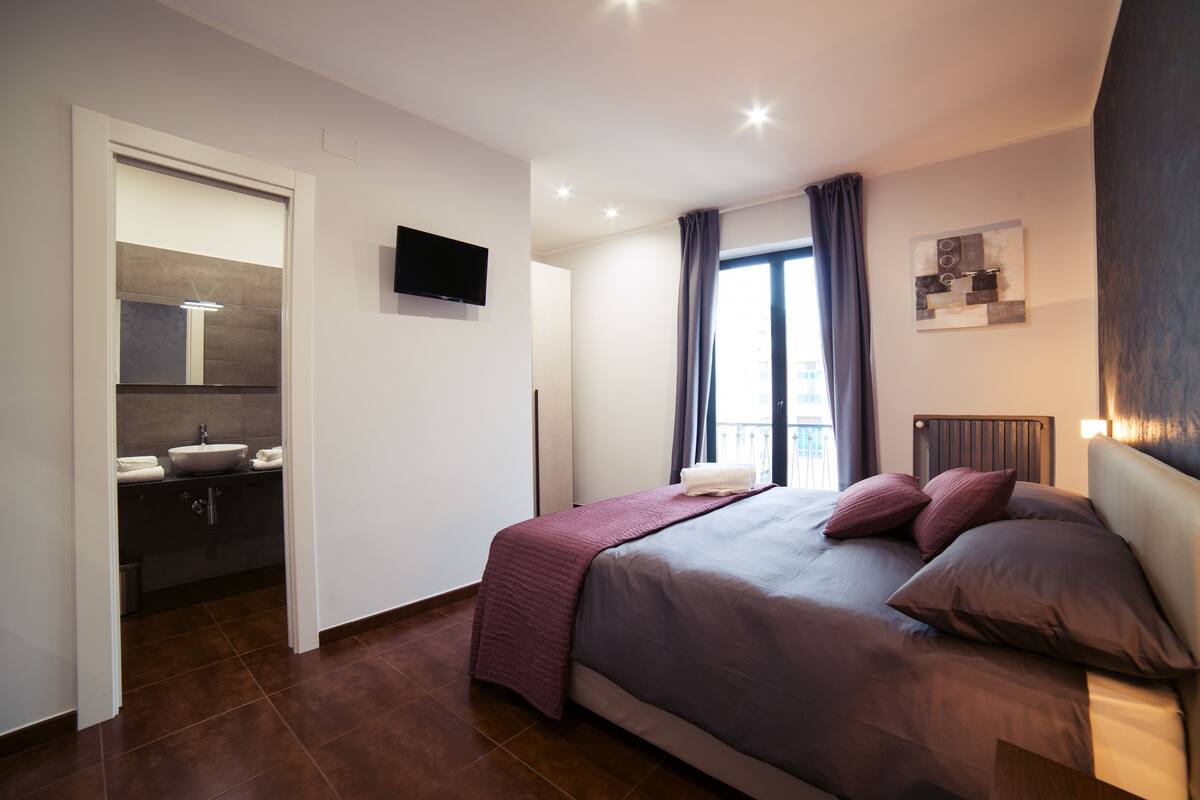 A modern hotel room with a bed, a TV, a bathroom with a sink, and a window with curtains.