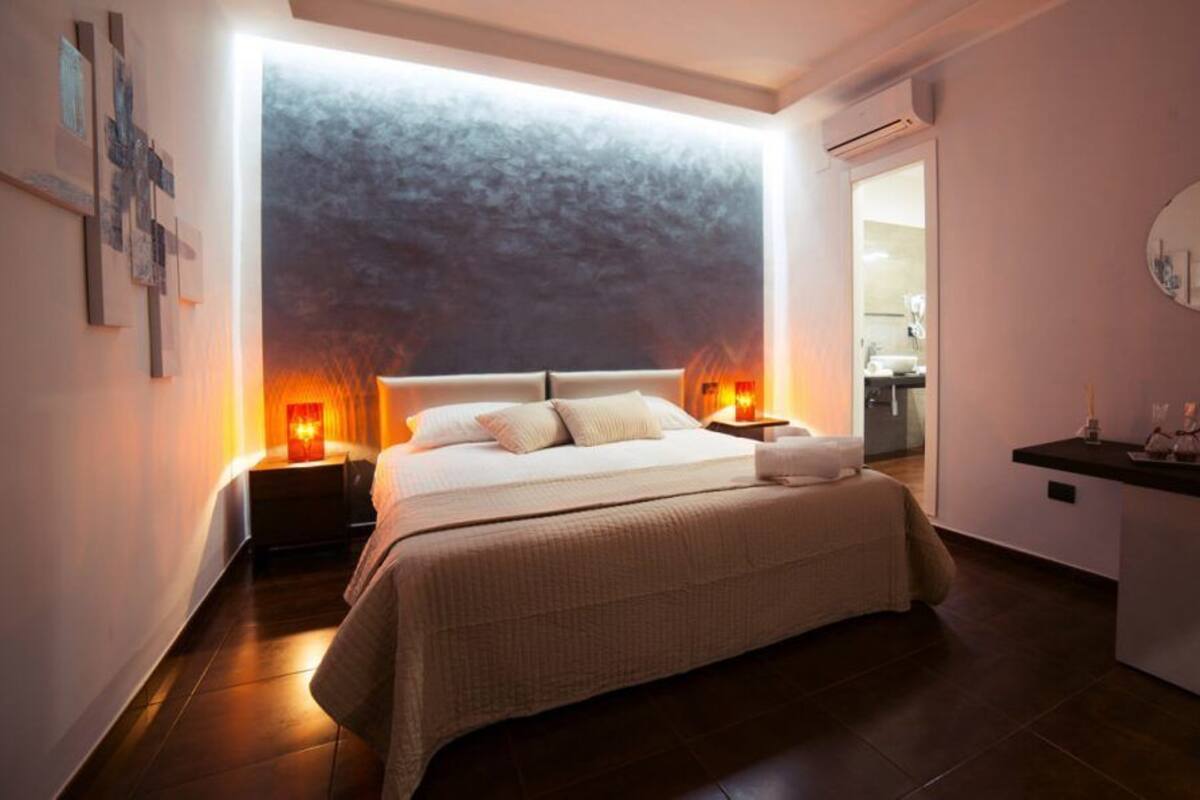 A modern bedroom with a large bed, bedside lamps, a small bedside table, and a wall-mounted mirror.