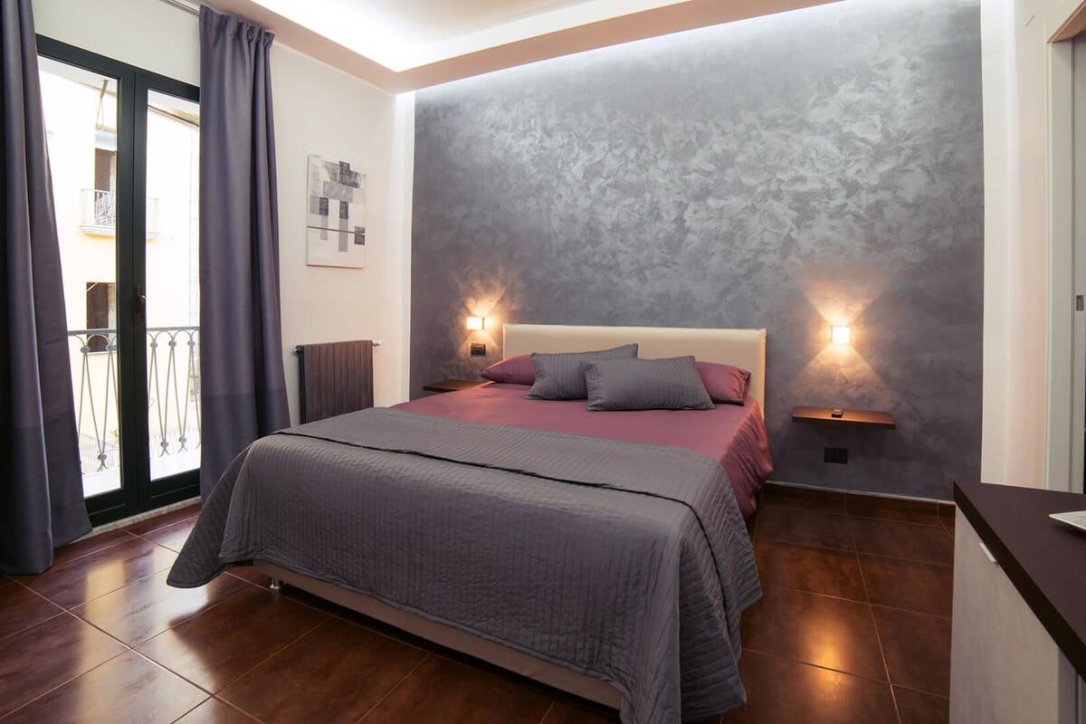 A bedroom with a large bed, two bedside lamps, a view of the balcony, and a grey textured wall.