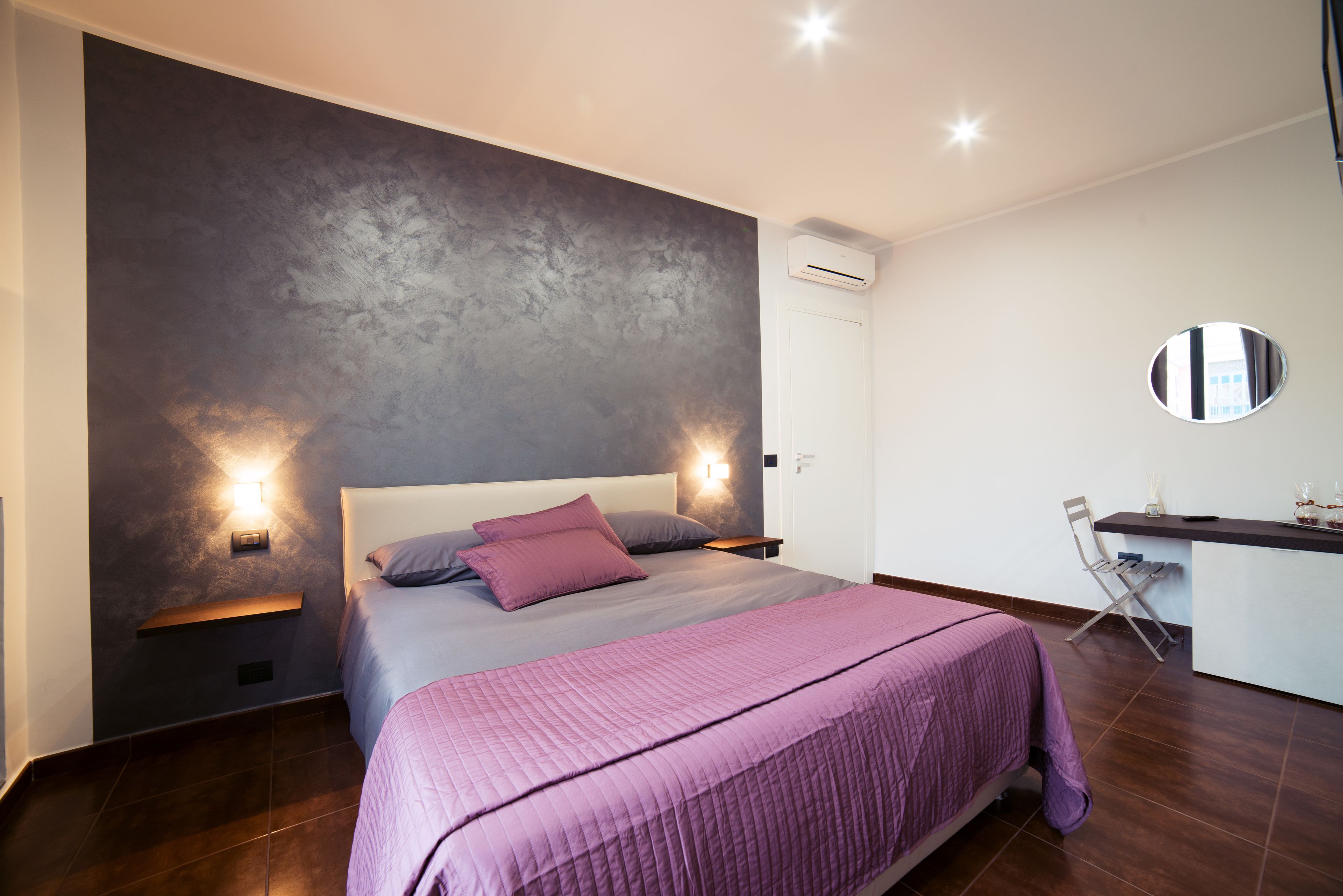 Un dormitorio moderno con una cama grande, una mesita de noche, un escritorio pequeño y un espejo redondo en la pared.