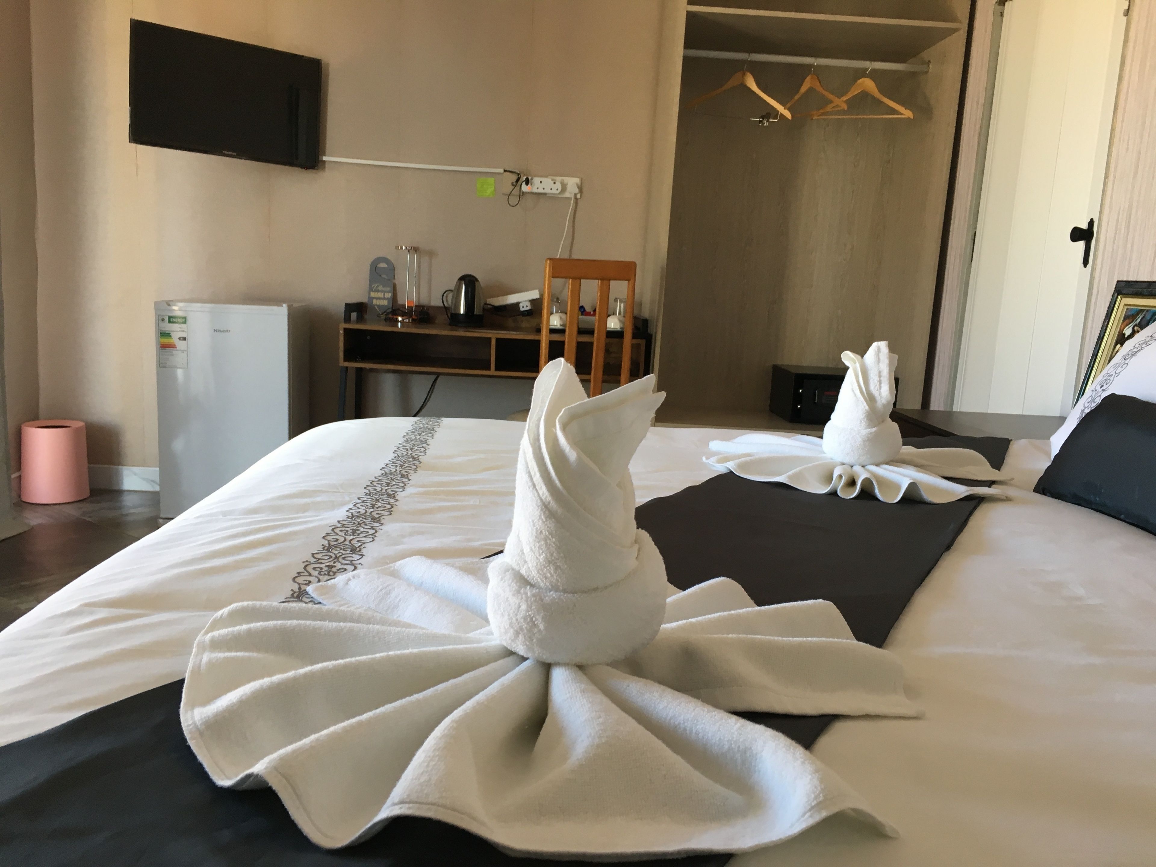 Habitación de hotel con una cama bien hecha, un televisor montado en la pared, un escritorio con una computadora y una lámpara, y un perchero con perchas.