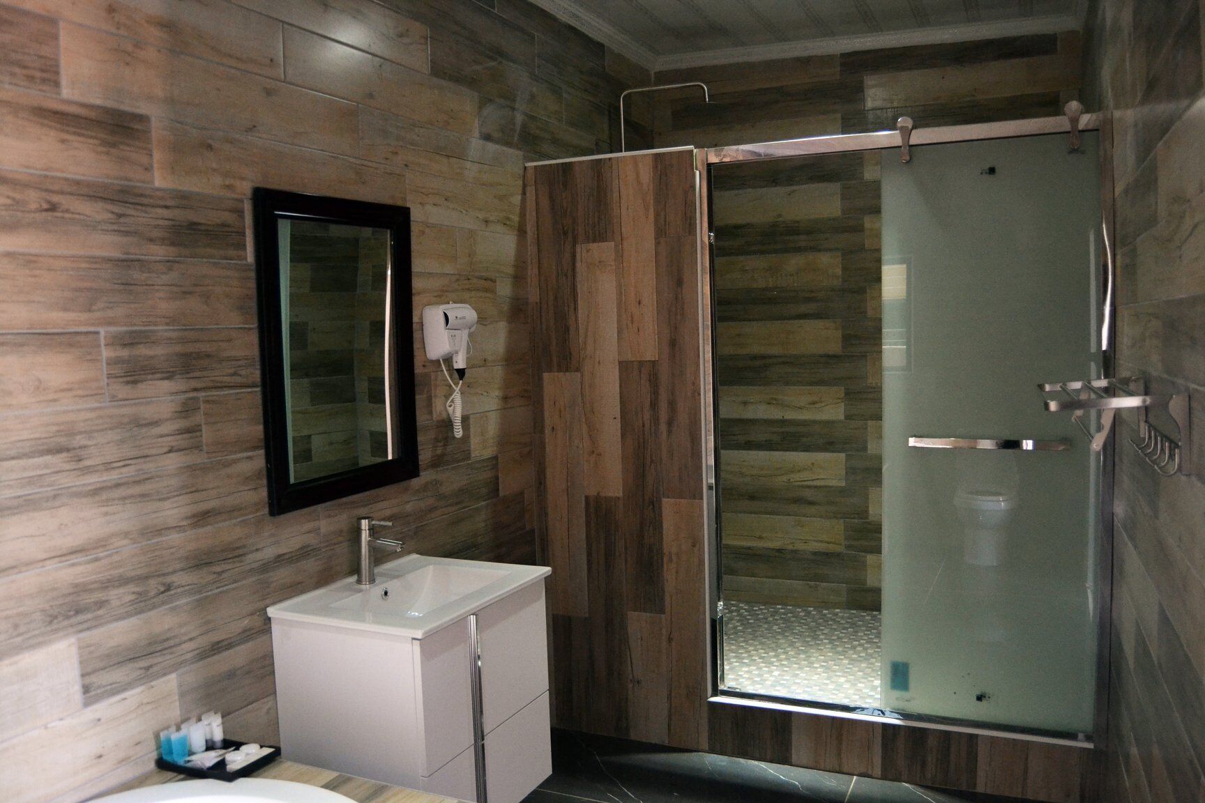 A modern bathroom with a wooden wall, a glass shower enclosure, a white sink, and a hairdryer mounted on the wall.