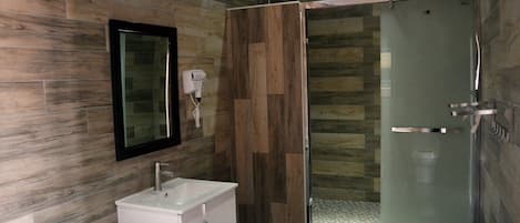 A modern bathroom with a wooden wall, a glass shower enclosure, a white sink, and a hairdryer mounted on the wall.