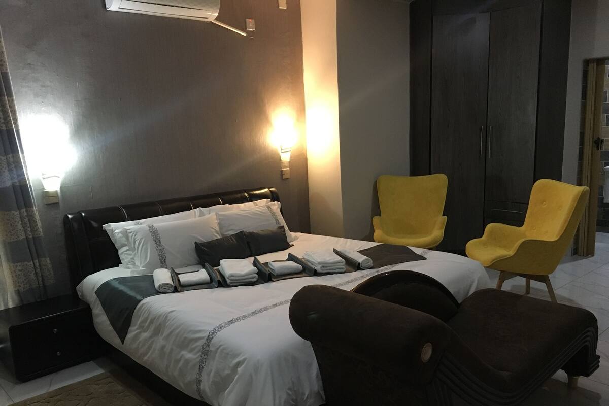 A hotel room with a bed, two yellow armchairs, a dark brown ottoman, and a wall-mounted air conditioner.