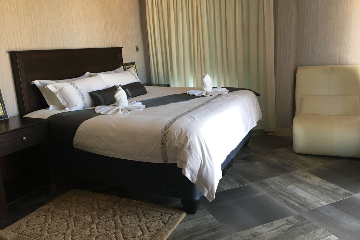 A neatly made bed with white and dark bedding, a wooden headboard, and a matching nightstand. A beige rug is placed in front of the bed, and a white armchair is positioned to the right.