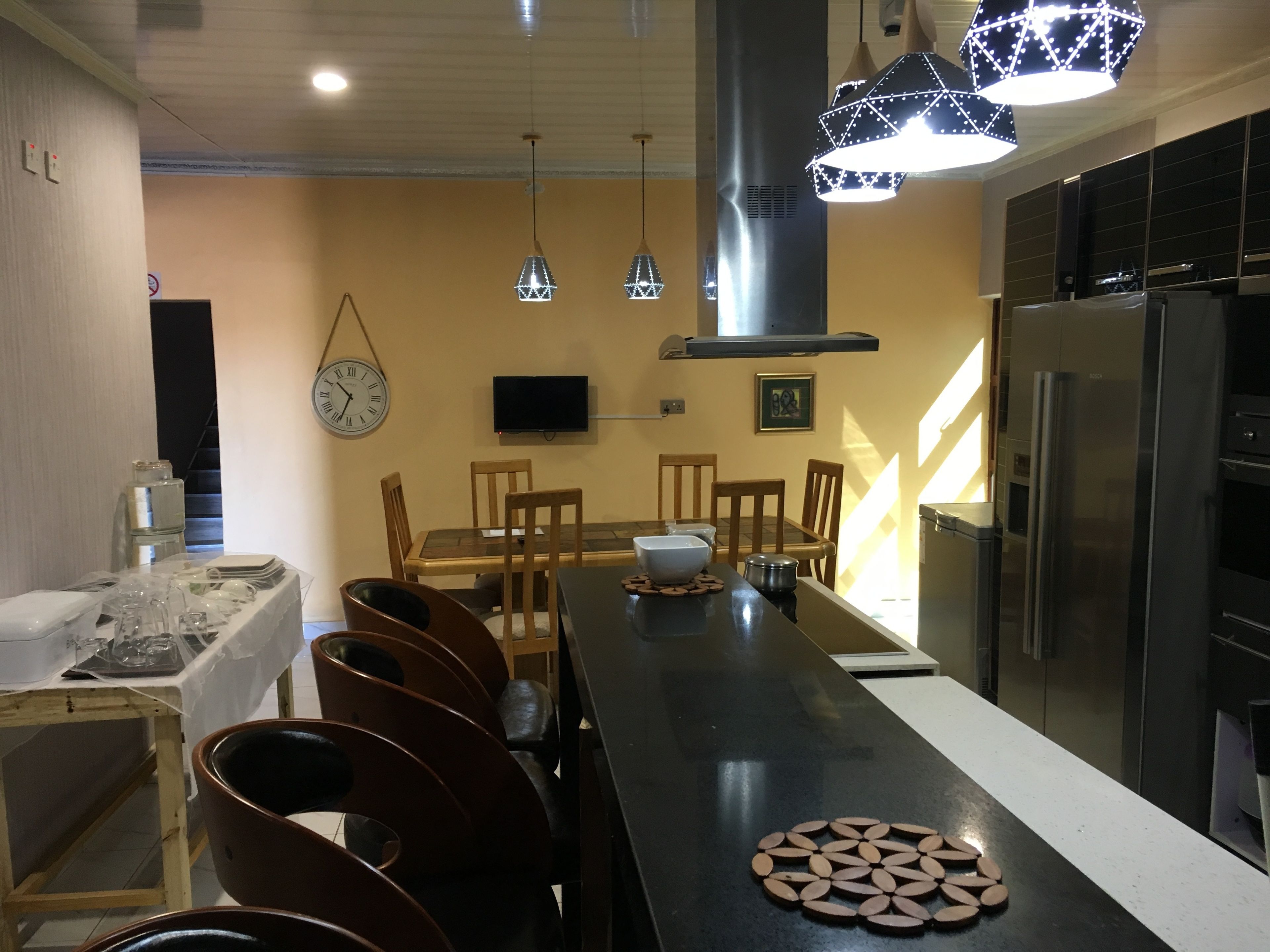 A modern kitchen with a long island, wooden chairs, a stainless steel refrigerator, and pendant lights.