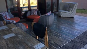 A sunlit outdoor seating area with wicker furniture and a cushioned chair, a wooden table, and a view of a swimming pool and buildings in the background. - Kanyi Guest House (Palapye)