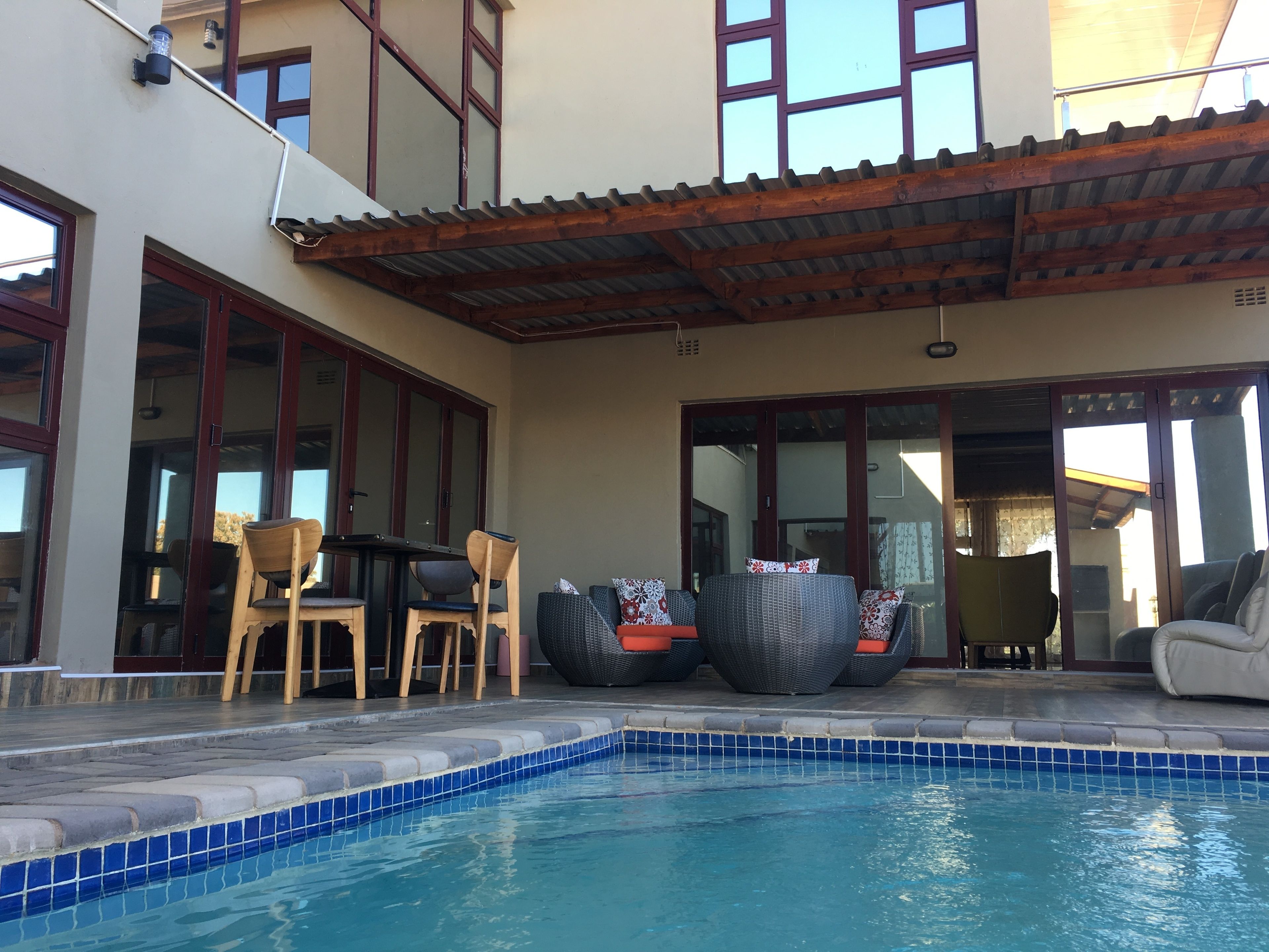 En swimmingpool med fliskanter, en overdækket terrasse med udendørs møbler og en bygning med store vinduer og et flistag.
