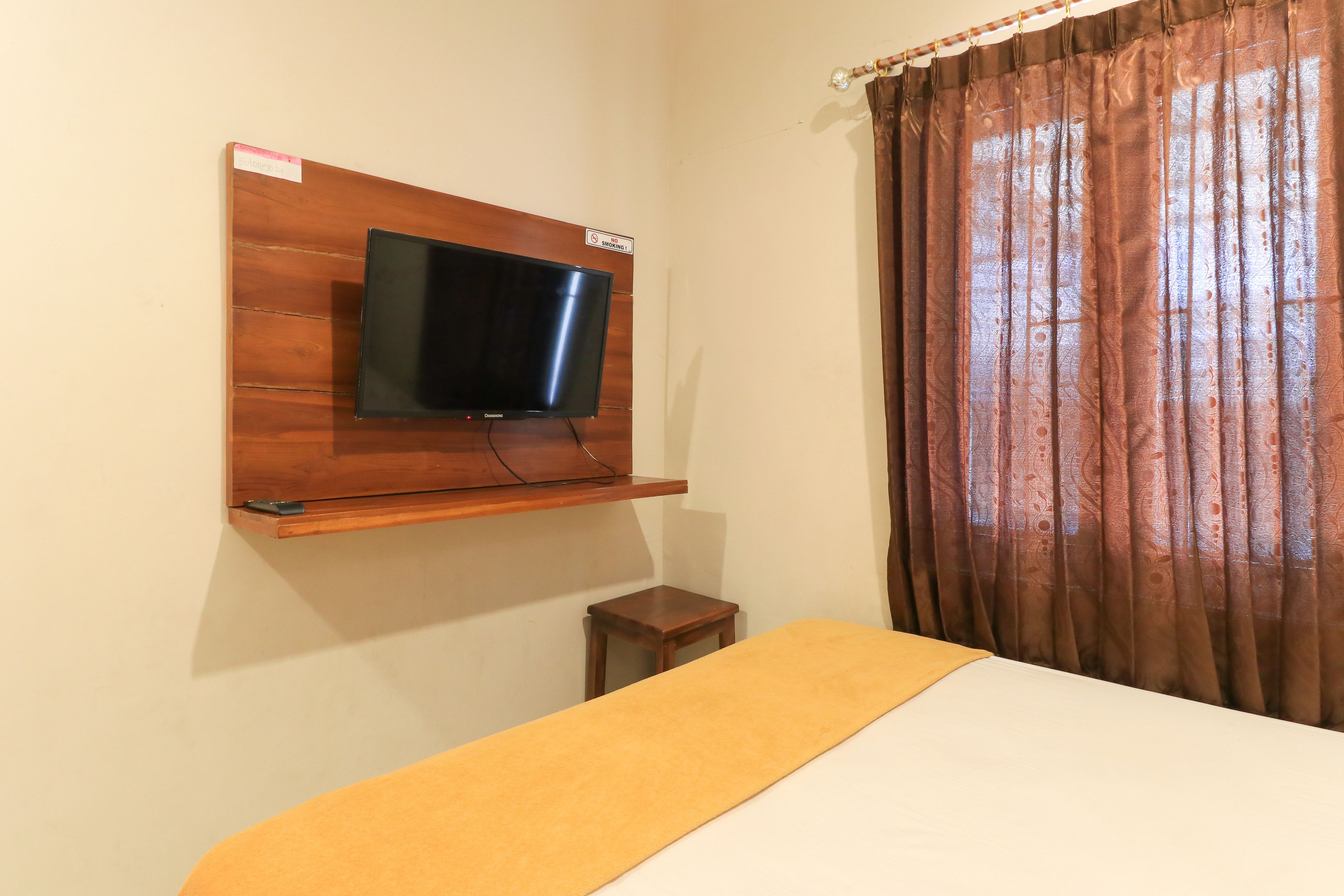 A room with a bed, a television mounted on a wooden panel, a small wooden stool, and a curtain.