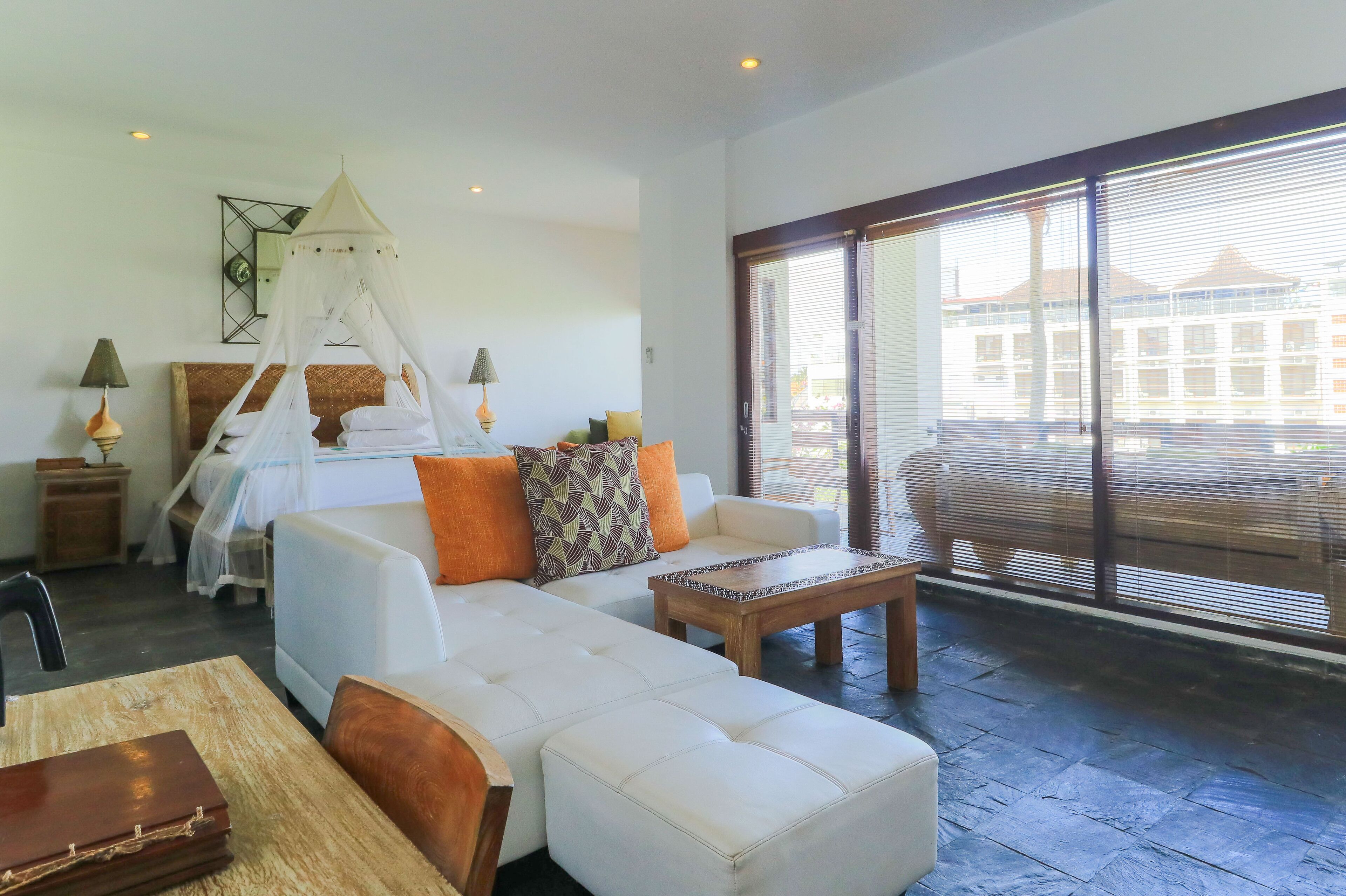 A modern living room with a white sofa, a wooden coffee table, and a large window with blinds.