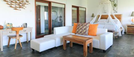 Una habitación de hotel moderna con un sofá blanco, una mesa de centro de madera, un escritorio con una silla y un ventanal grande.