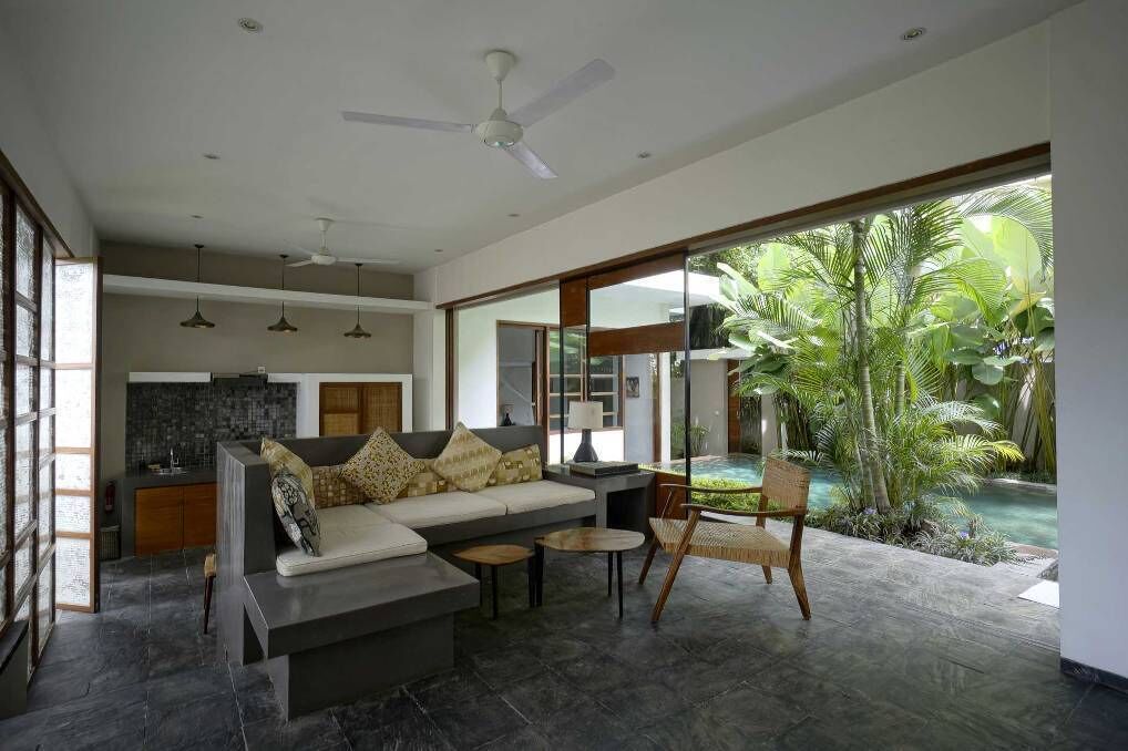 Ein modernes Wohnzimmer mit Sofa, Couchtisch und Sessel, im Hintergrund eine Küche und Blick auf einen Swimmingpool sowie tropische Pflanzen.