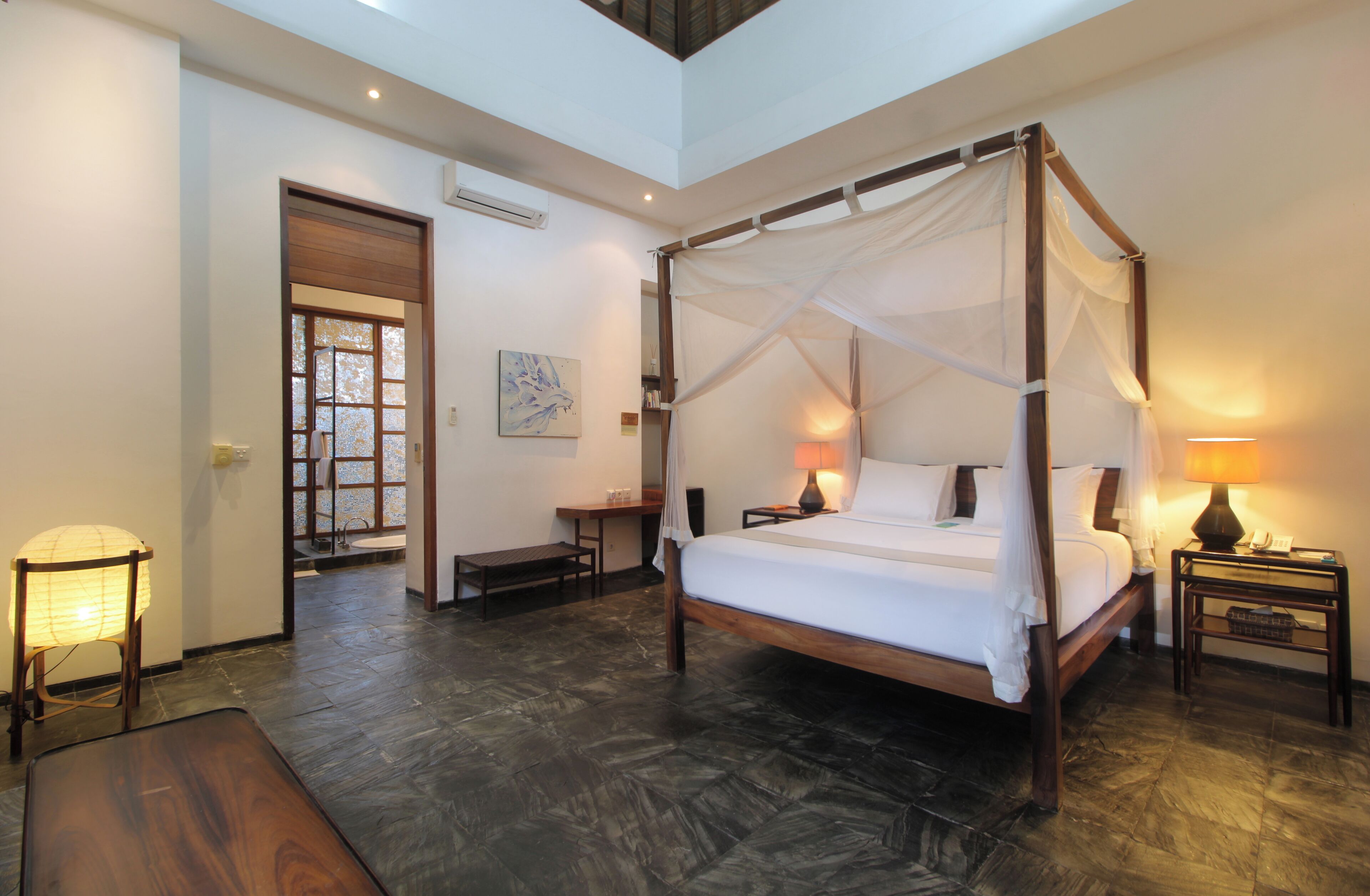 A four-poster bed with a canopy, bedside tables with lamps, a wooden bench, and a view of the outside through a door.