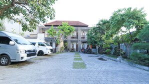 A paved courtyard with parked vehicles, a building with a tiled roof, and surrounding greenery. - Hotel Warta Putra (Denpasar)