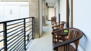 A corridor with a wooden railing, a dining area with wooden chairs, and a view of a building with a red roof in the background. - Hotel Warta Putra (Denpasar)