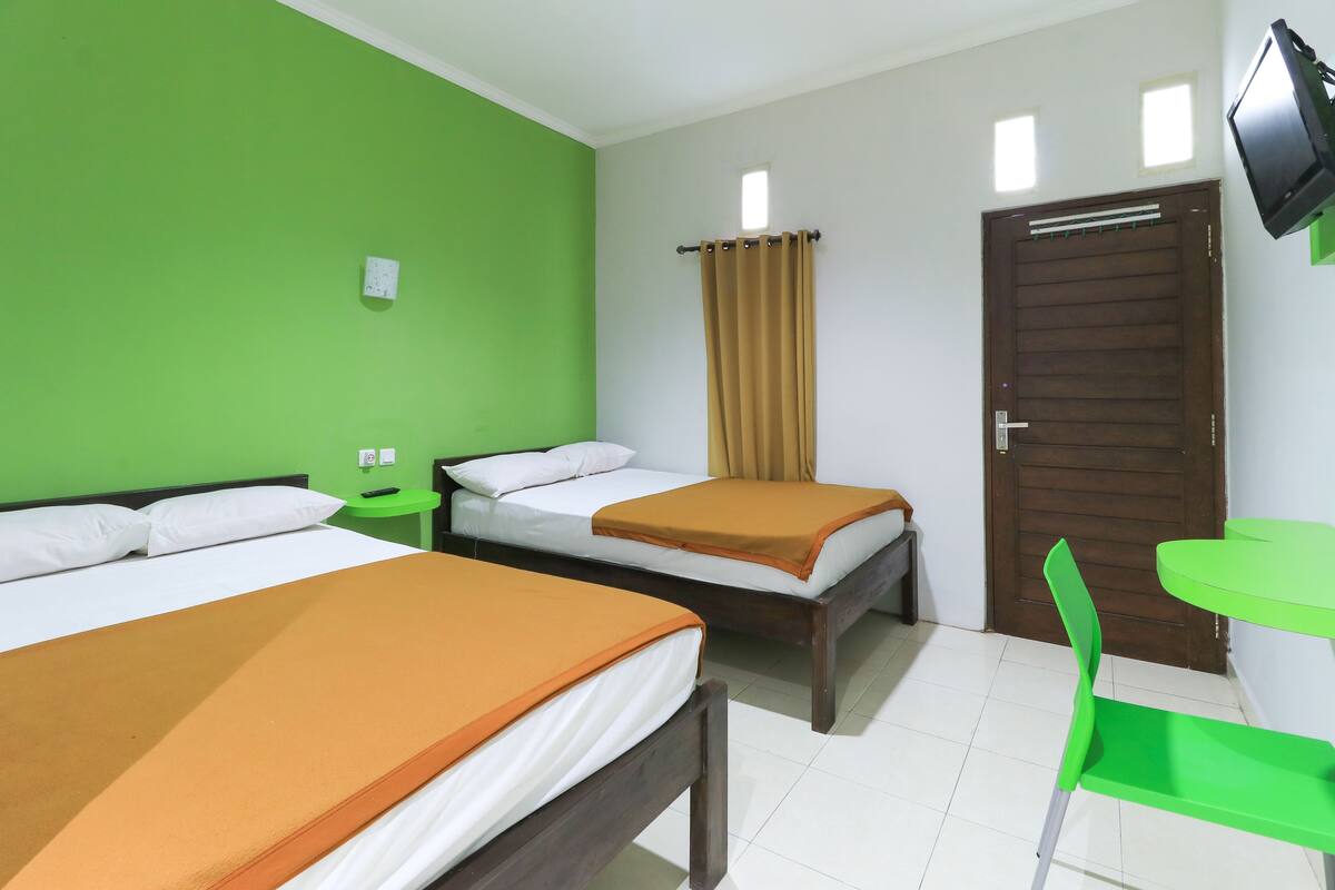 A hotel room with two beds, a green chair, a small table, a TV mounted on the wall, and a door with a curtain.
