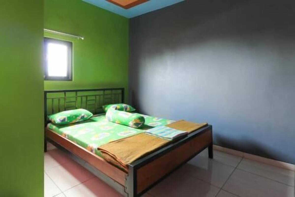 A single bed with a green and white patterned bedspread, a wooden headboard, and a window with a white blind.