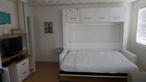 A bedroom with a bed, a TV on a shelf, and a window with blinds. - GROUND FLOOR STUDIO ~ BEACHSIDE condo with a kitchen, walkout to pool.FN #401 (St Pete Beach)